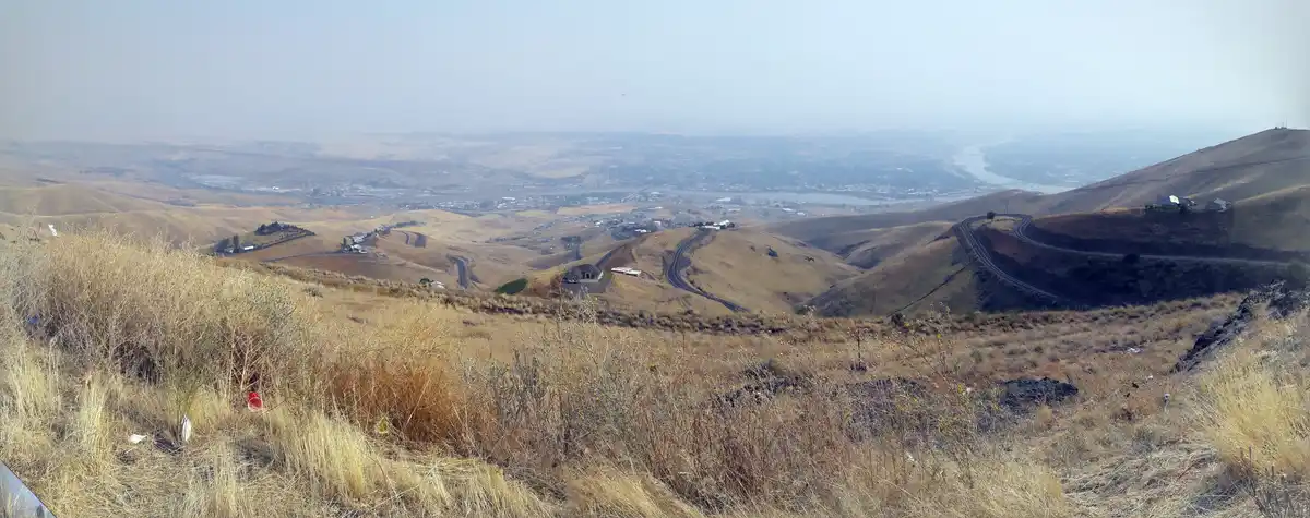 A panoramic view from a dry, grassy hill overlooking a valley with winding roads, distant towns, and a river, with overlaid…