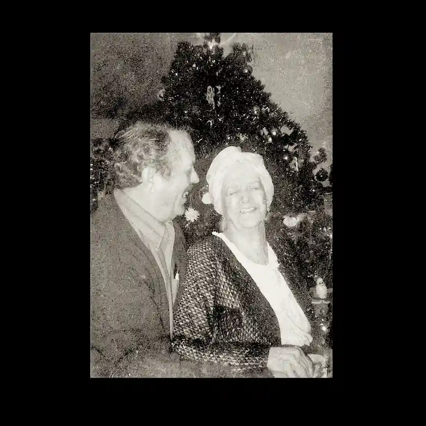 A grainy black-and-white photo shows an older man and woman smiling and looking at each other in front of a decorated…