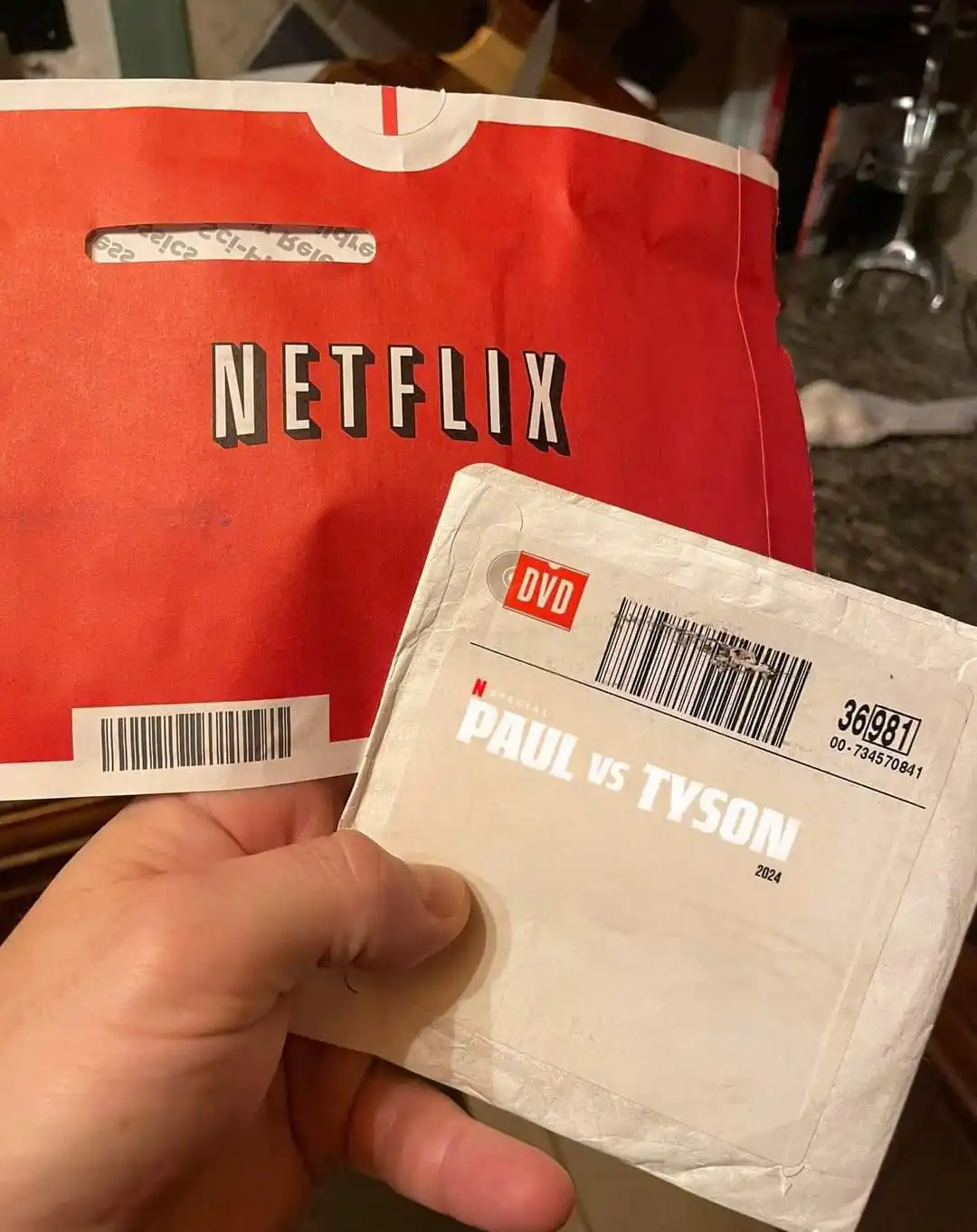A hand holds a white DVD case labeled 'N SPECIAL PAUL VS TYSON 2024' in front of a red Netflix bag. The DVD case has a…