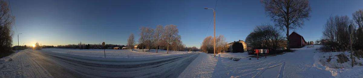 A wide-angle winter panorama shows a snow-covered rural road curving through a quiet landscape with bare trees, houses, and…