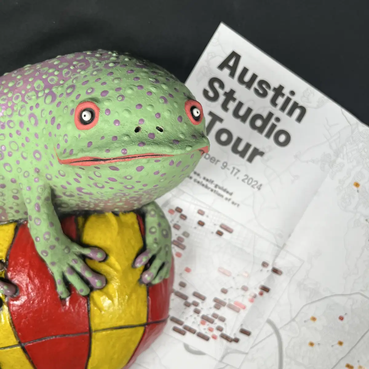 A green frog statue with purple spots and red-rimmed eyes sits atop a red and yellow tiled object, next to a printed map for…
