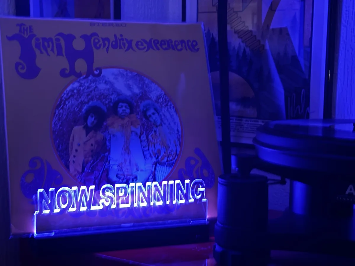 A vinyl record album cover for 'The Jimi Hendrix Experience' is displayed next to a record player under blue lighting. A…