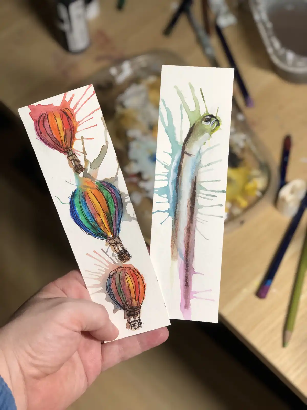 A hand holds two watercolor paintings on small rectangular cards. One card shows three colorful hot air balloons with…