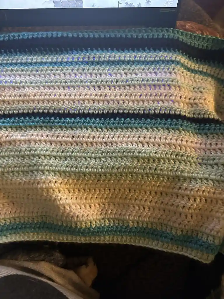A close-up photo shows a hand-crocheted blanket with horizontal stripes of teal, cream, and light green yarn draped over a…