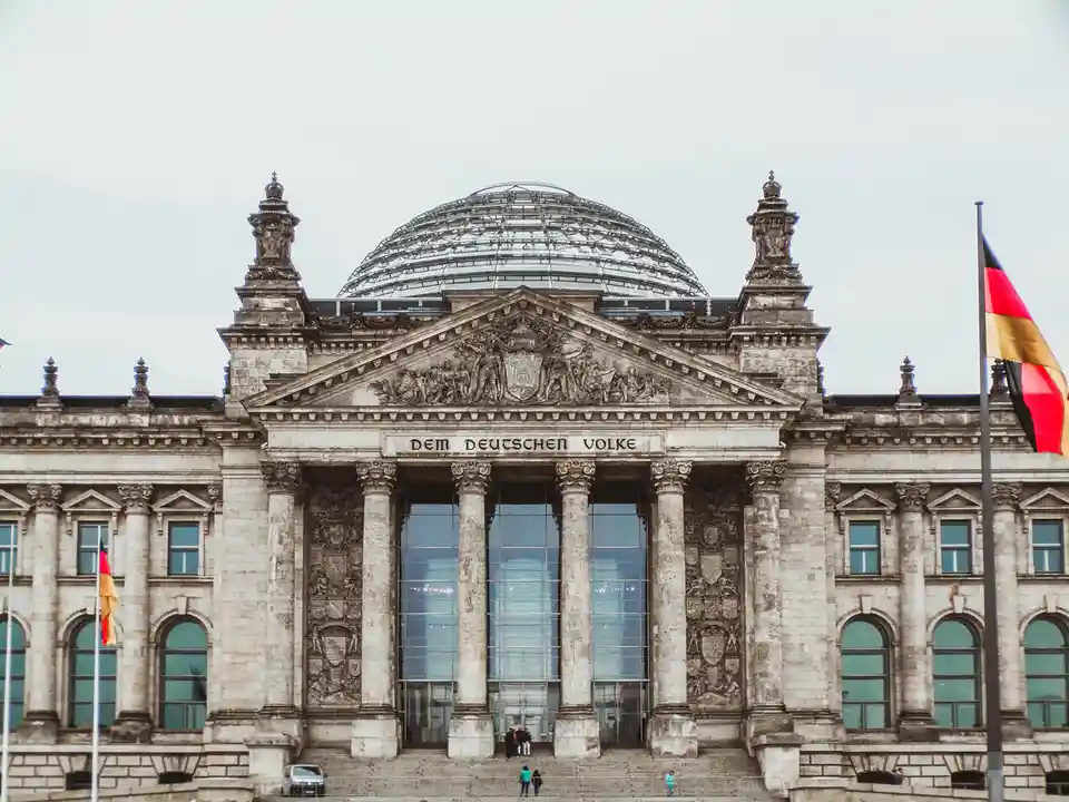 The image shows the Reichstag building in Berlin, Germany, featuring its iconic glass dome and classical stone facade with…