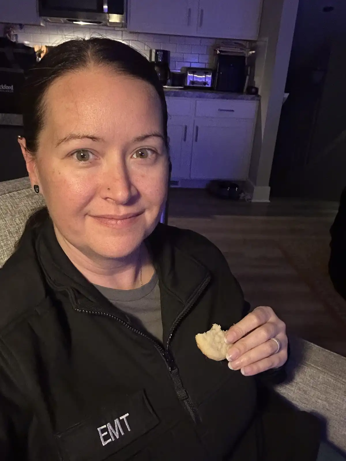 A woman wearing a black jacket with 'EMIT' embroidered on the chest sits in a dimly lit room, holding a partially eaten…
