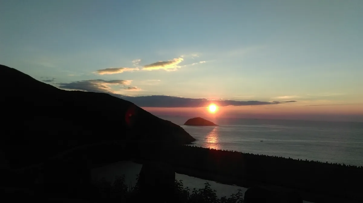 A serene sunset over the ocean with a silhouetted hill on the left and a small island in the distance. The sun is low on the…