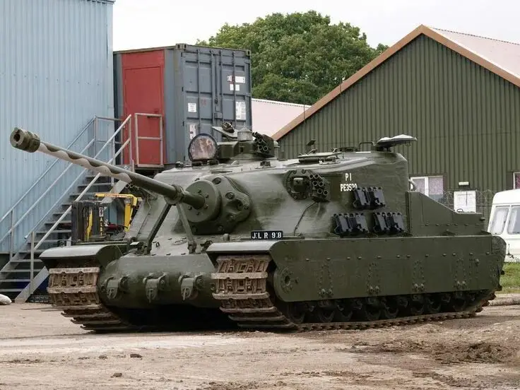 A green military tank with the text 'PI PESSON' painted on its side is parked outdoors near industrial buildings and…