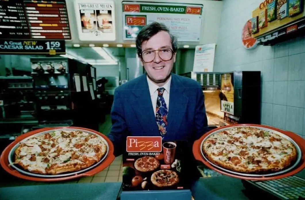A man in a suit and glasses stands behind a counter in a fast-food restaurant, holding two large pizzas on red trays. In…