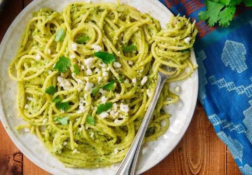 a plate of spaghetti coated in green pesto sauce, topped with crumbled white cheese and fresh cilantro leaves, with a fork…