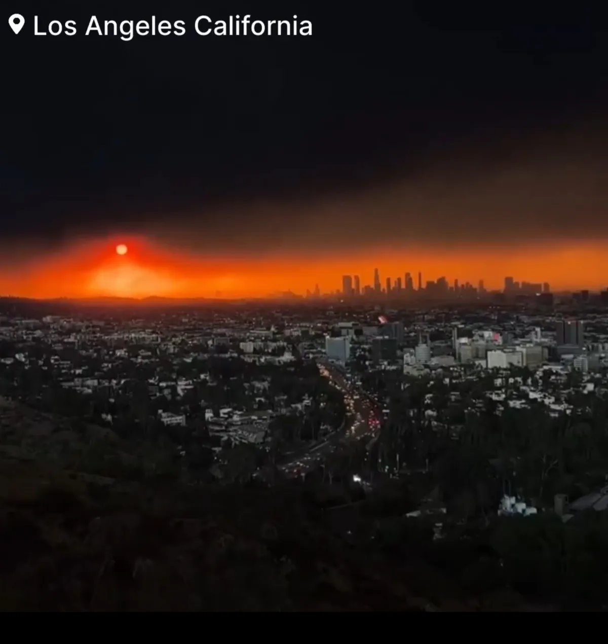 An aerial view of Los Angeles at dusk or dawn, where the sky is choked with thick orange and black smoke from wildfires…