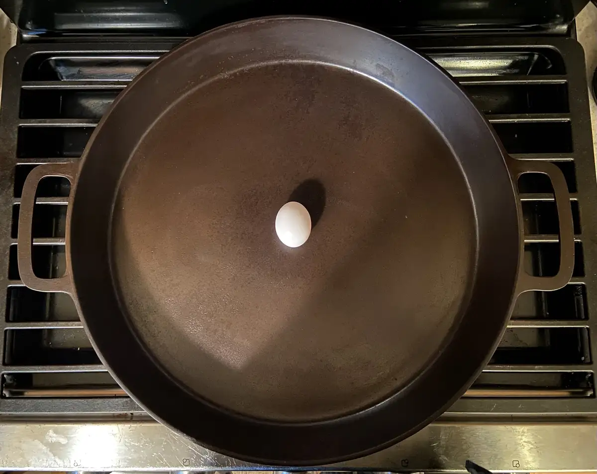 A single white egg sits centered in a large, empty, dark brown cast iron skillet placed on a gas stove burner. The image is…