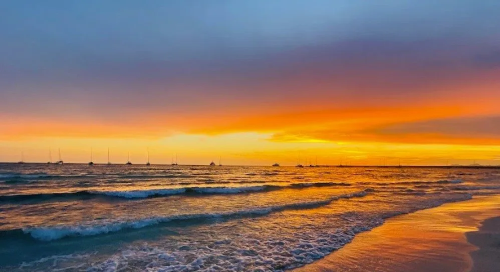 A serene beach sunset with orange and purple skies, waves rolling onto shore, and sailboats silhouetted on the horizon…