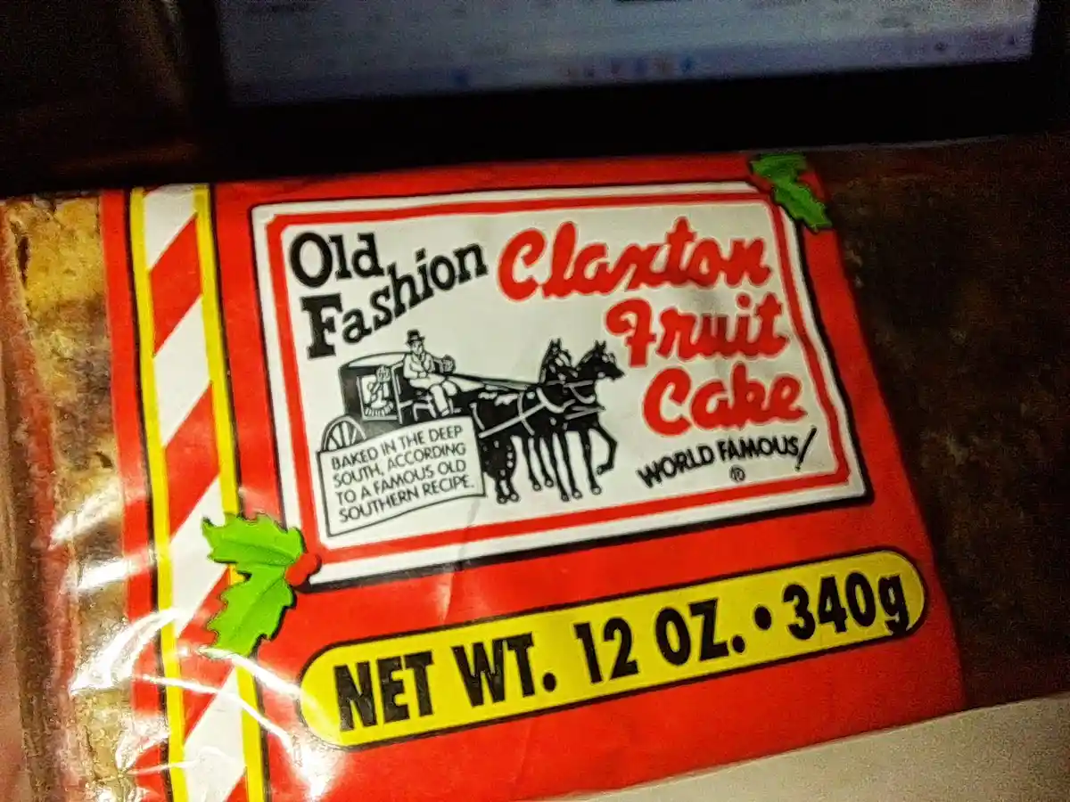 A close-up of a red-wrapped Claxton Fruit Cake box featuring a vintage illustration of a man in a carriage pulled by two…