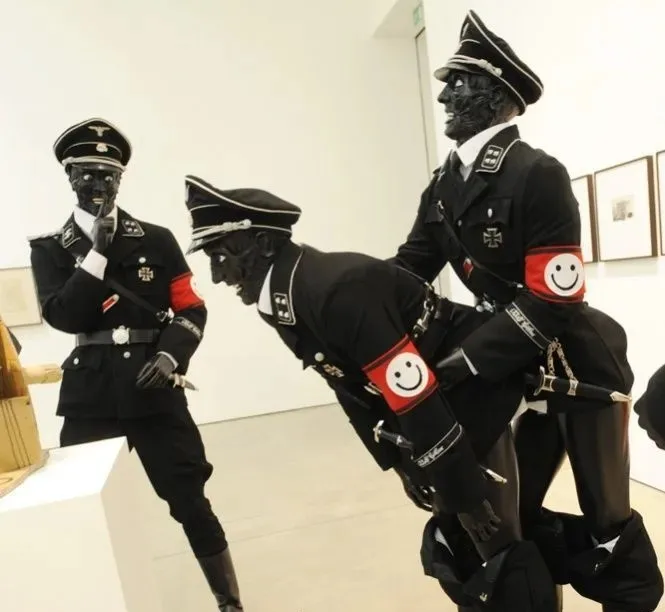 Three black-painted figures in Nazi uniforms with smiley-face armbands are posed in a gallery; one is bent over another…