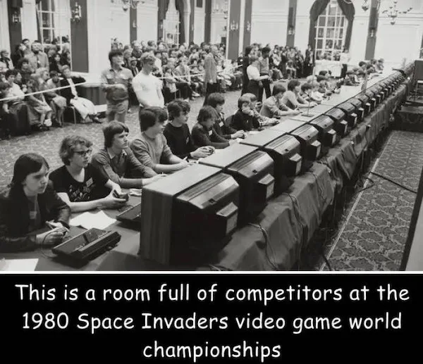 A black-and-white photo shows a long table in a large room filled with children and teenagers seated in front of bulky…