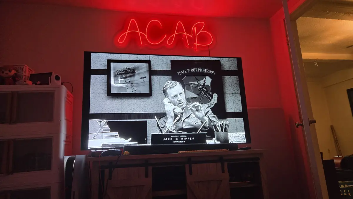 A red neon sign spelling 'ACAB' is mounted above a television displaying a black-and-white scene from the movie 'Dr…