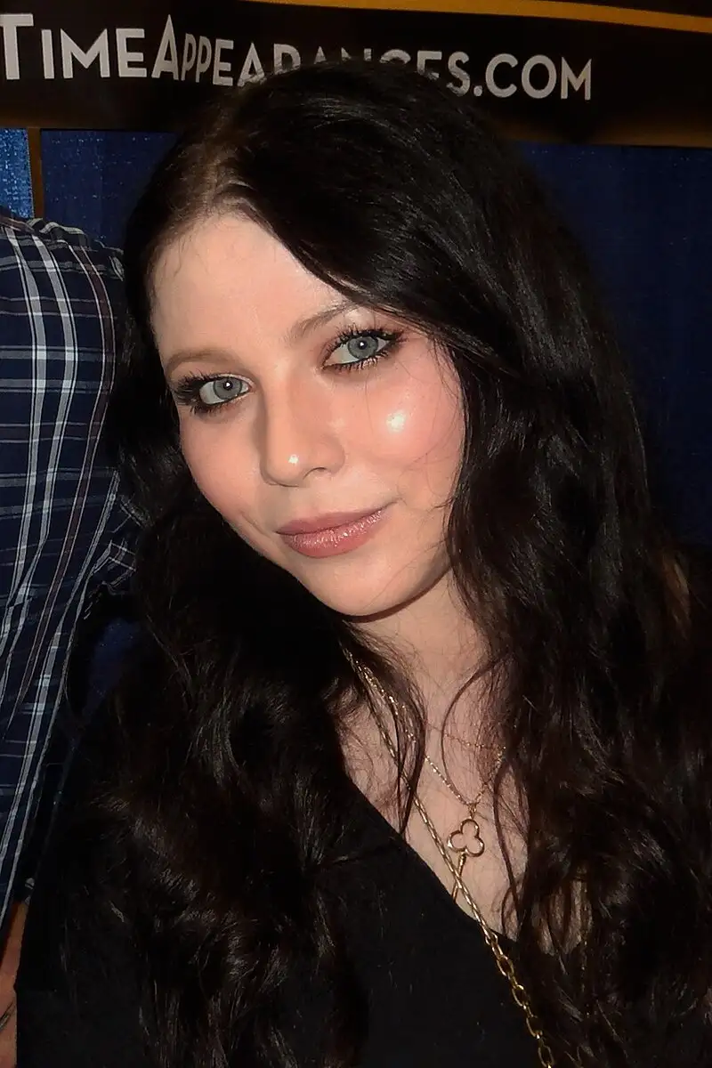 A close-up photo of a woman with long dark wavy hair, light-colored eyes, and a subtle smile, wearing a black top and gold…
