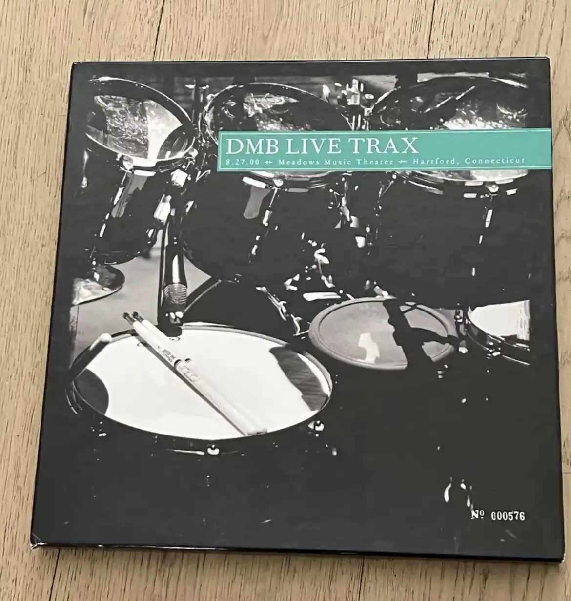 A black and white photo of a drum set with drumsticks resting on a snare drum, overlaid with the text 'DMB LIVE TRAX 8.27.00…