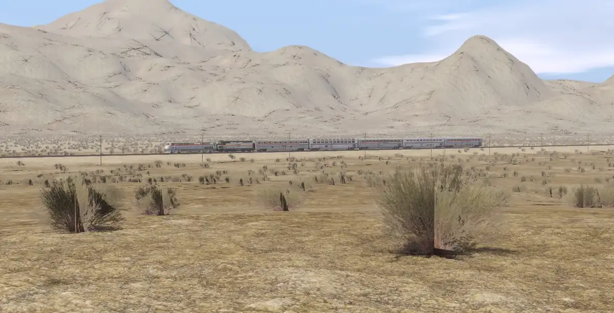 A long silver passenger train travels across a vast, arid desert landscape with sparse shrubs in the foreground and large…