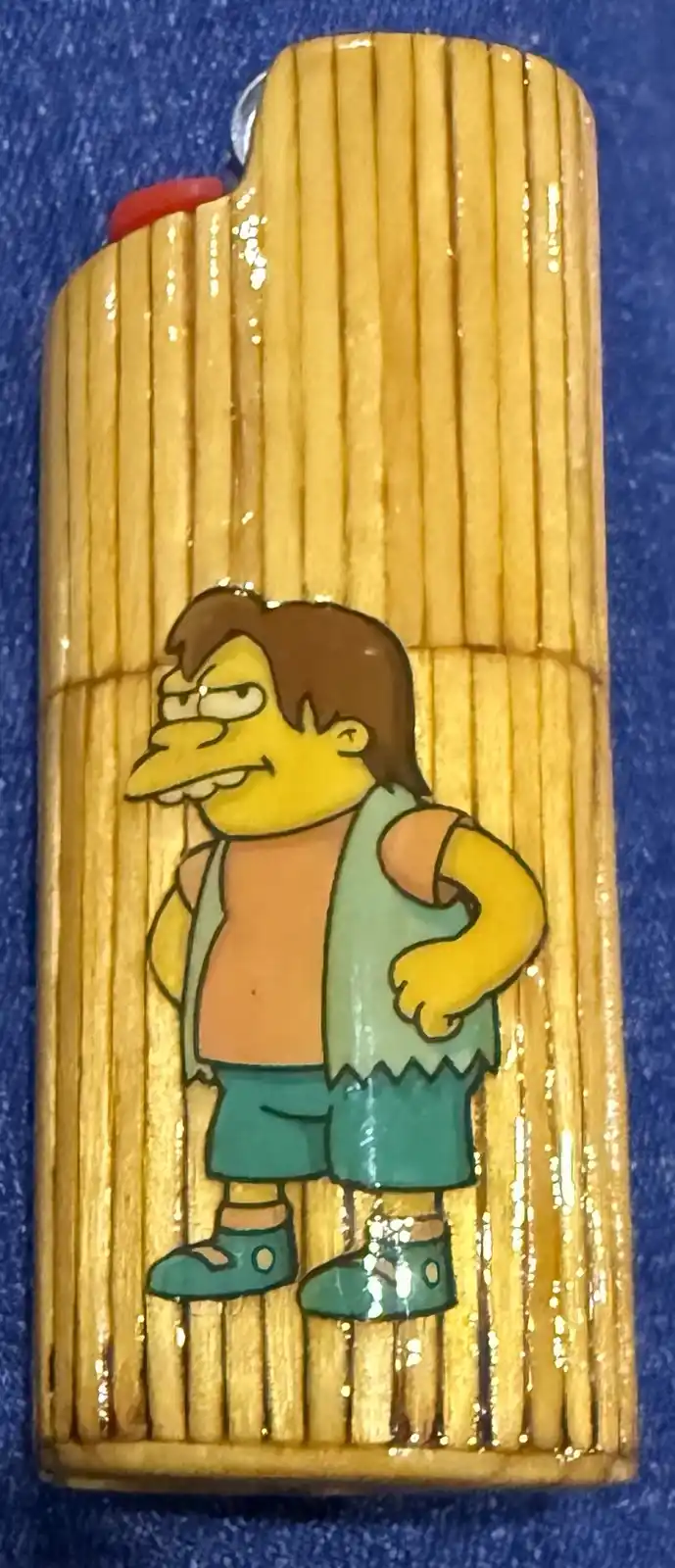 A close-up photo of a yellow bamboo-textured lighter with a cartoon character printed on it. The character is Homer Simpson…