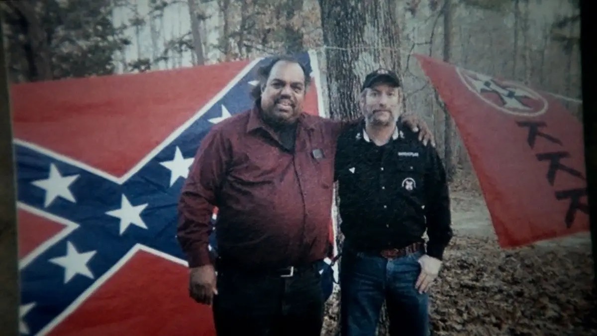 Two men stand together outdoors in front of two large flags: a Confederate flag on the left and a red flag with a white…
