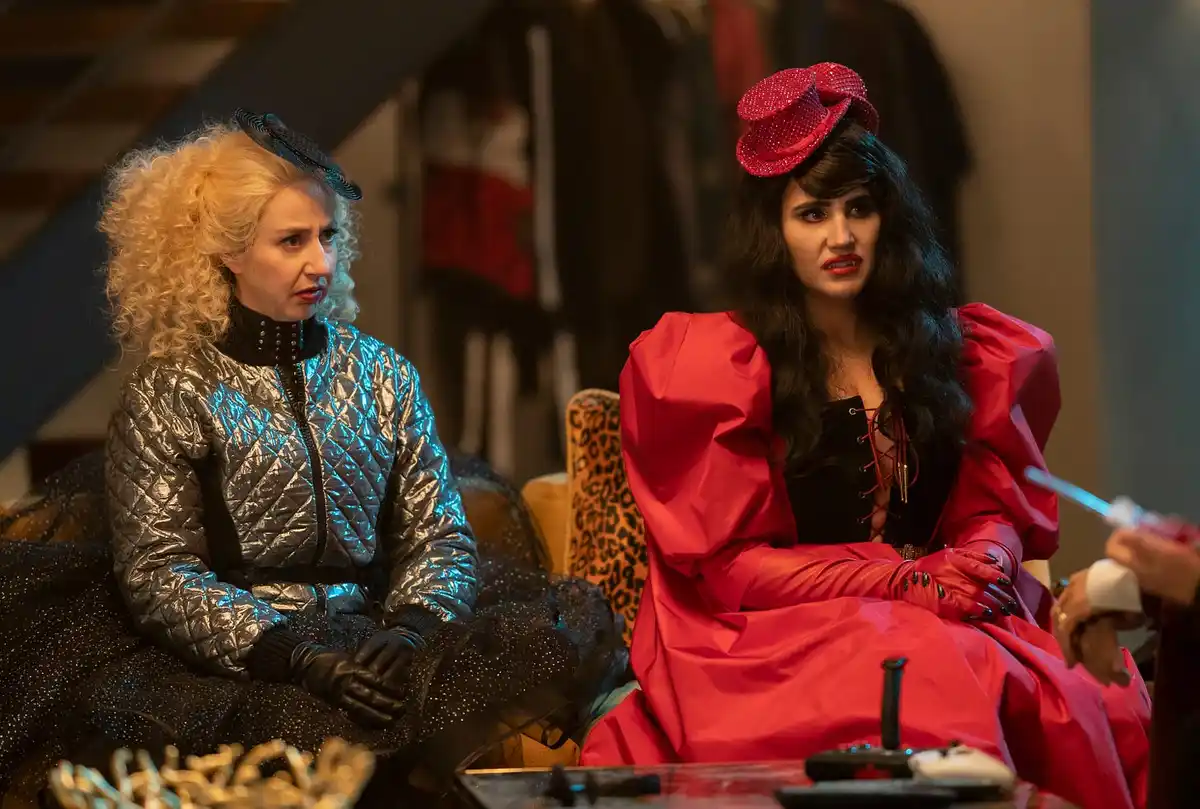 two women in elaborate costumes sit on a couch; one in a silver quilted jacket and black gloves, the other in a red dress…
