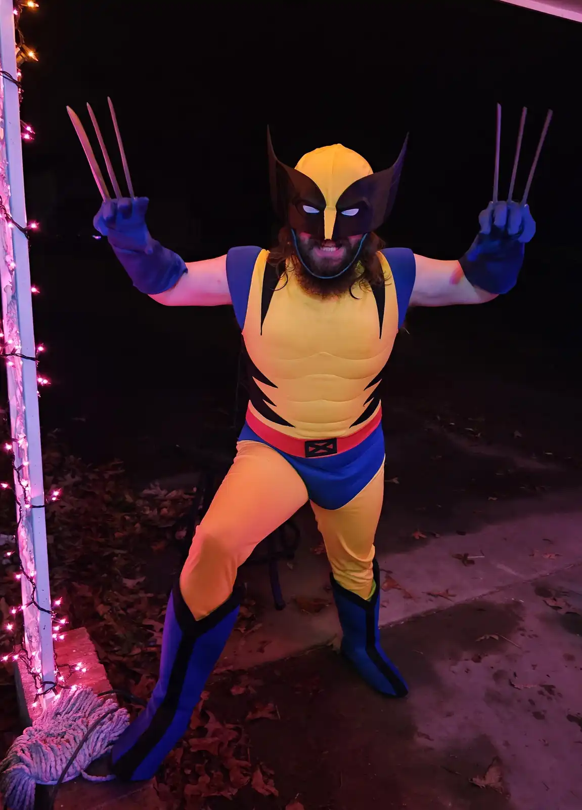 A person in a full-body Wolverine costume, complete with mask, claws, and yellow-and-blue suit, poses outdoors at night with…