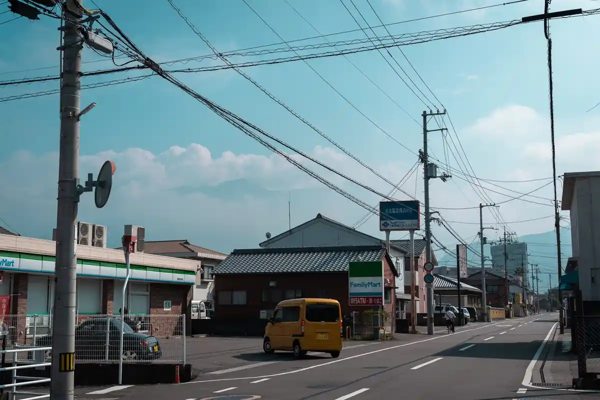 A street scene in Japan with a yellow van driving past a FamilyMart convenience store, power lines crisscrossing the sky…