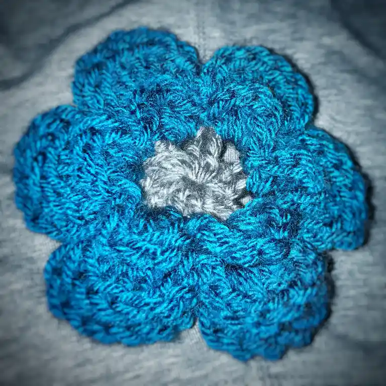 A close-up photo shows a hand-crocheted flower made of bright blue yarn with a gray center, attached to a gray fabric…