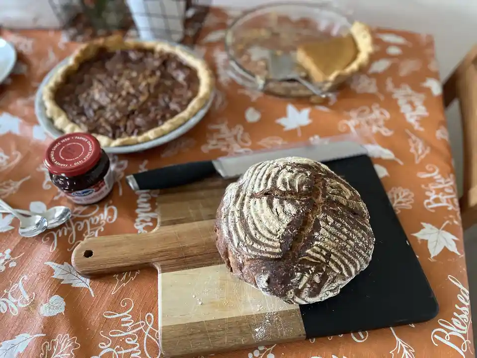 A rustic, round loaf of artisan bread with a scored top sits on a wooden and black cutting board, placed on an orange…