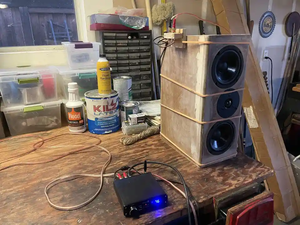 A homemade speaker cabinet made of three stacked wooden boxes, bound with orange straps, sits on a cluttered workbench next…