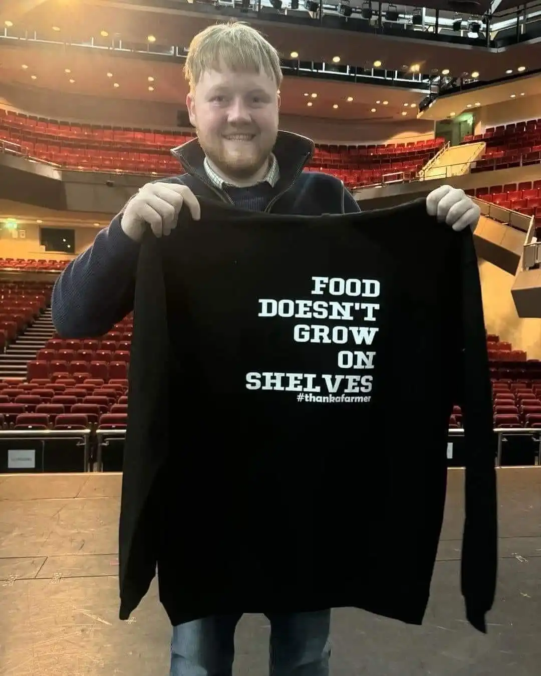 A smiling man with blond hair and a beard stands on a stage in an empty theater, holding up a black sweatshirt. The…