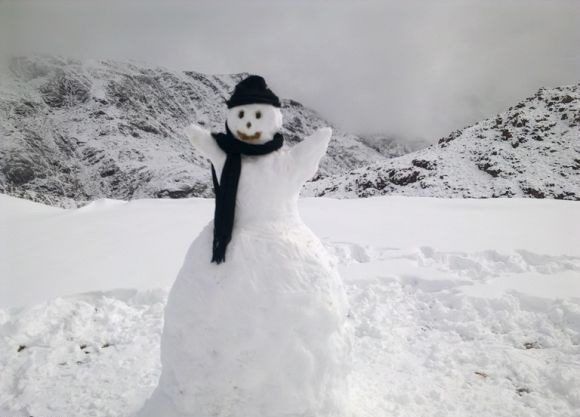 A snowman stands in a snowy mountain landscape, wearing a black beanie and scarf, with arms raised. The overlaid text reads…