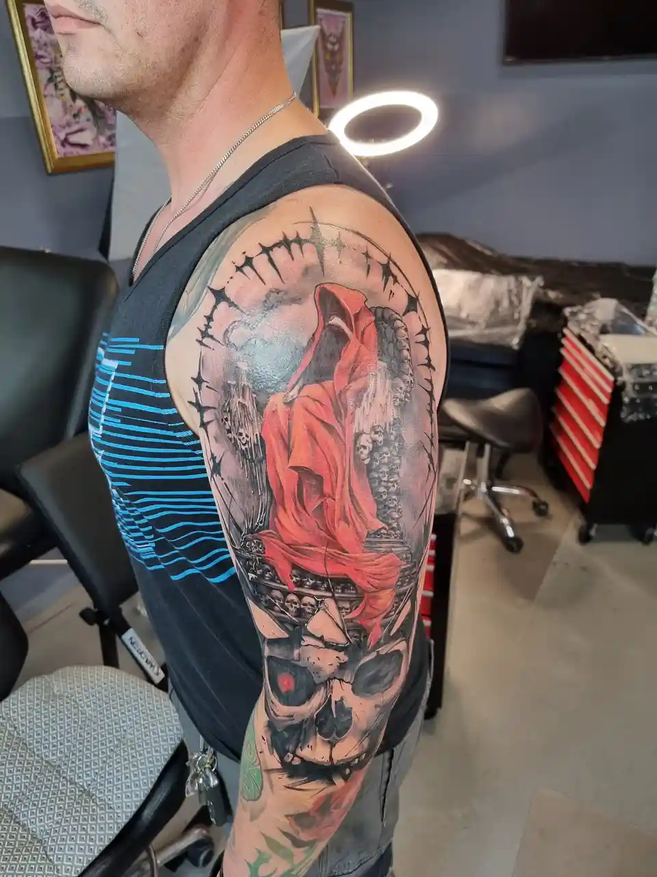 A man in a tattoo parlor shows off a large, detailed sleeve tattoo on his left arm featuring a grim reaper in a red cloak…