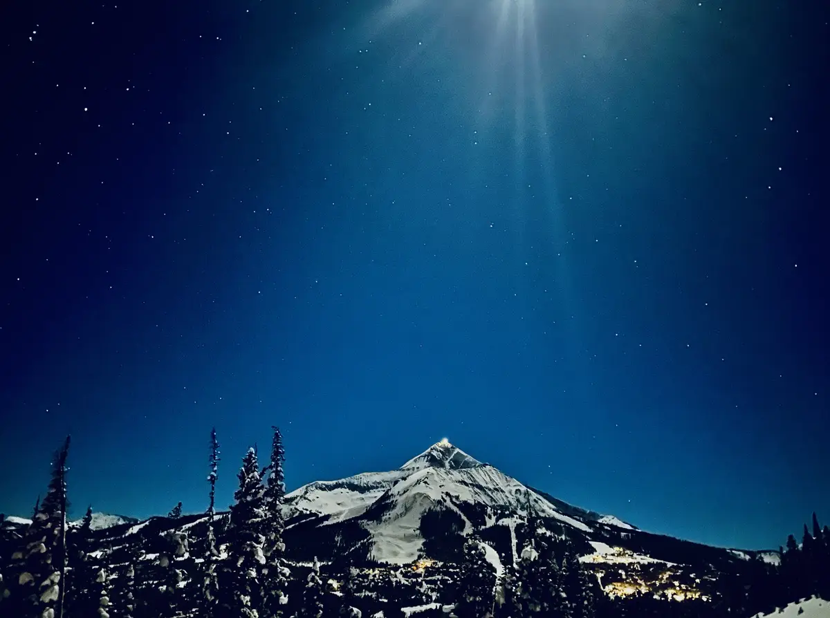 A serene night photograph of a snow-capped mountain under a starry sky with a bright beam of light descending from above…