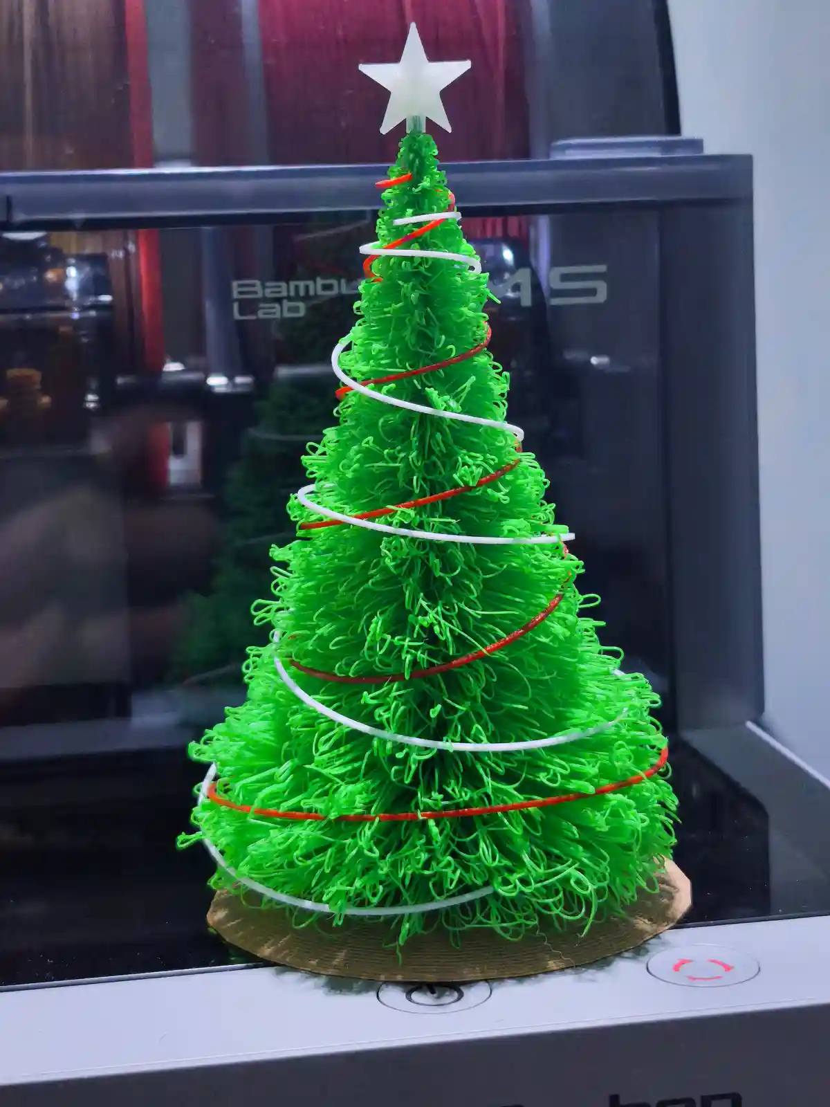 A small, bright green 3d-printed Christmas tree with red and white spiral garlands and a white star on top sits on a golden…