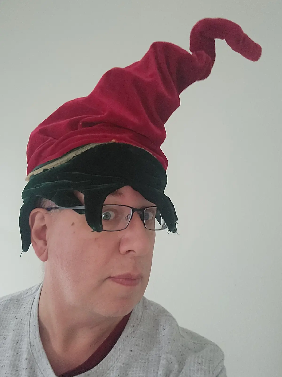 A person wearing a red velvet jester hat with a black blindfold over their eyes, glasses, and a grey shirt, looking…