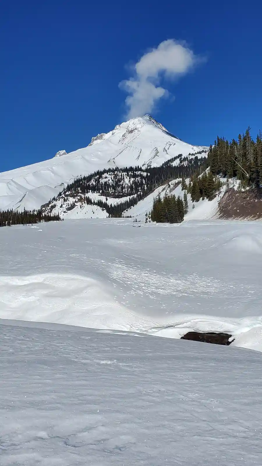A snow-covered mountain under a clear blue sky with a single, distinct cloud directly above its peak, shaped like a question…