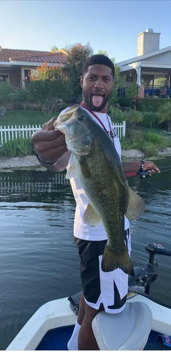a man stands on a boat holding a largemouth bass, sticking his tongue out in a playful, exaggerated manner. He wears a white…