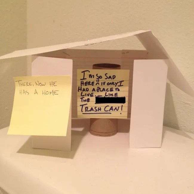 A handmade cardboard house with two sticky notes. One yellow note on the left reads 'THERE, NOW HE HAS A HOME'. The other…