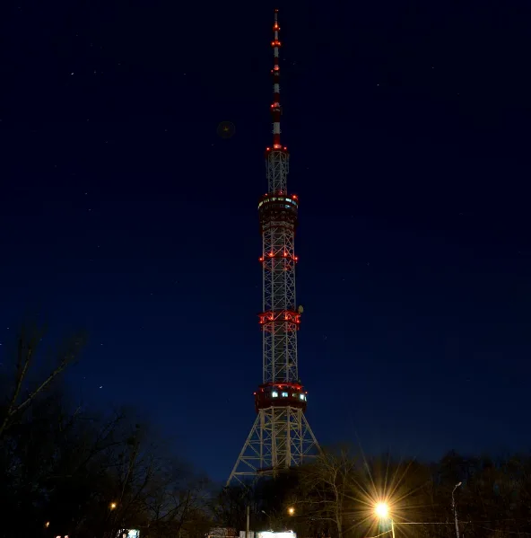 A tall, illuminated lattice tower with red lights stands against a dark night sky filled with stars. A small, faint moon is…