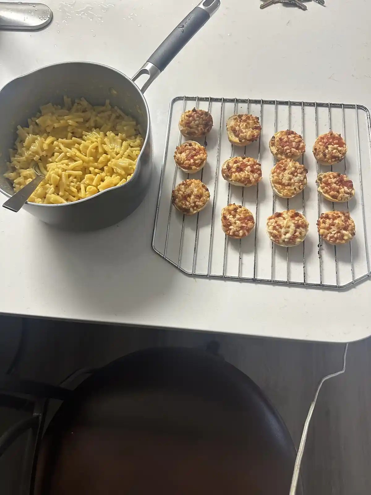 A kitchen counter shows a pot of mac and cheese next to a wire rack holding twelve mini pizzas made with English muffin…