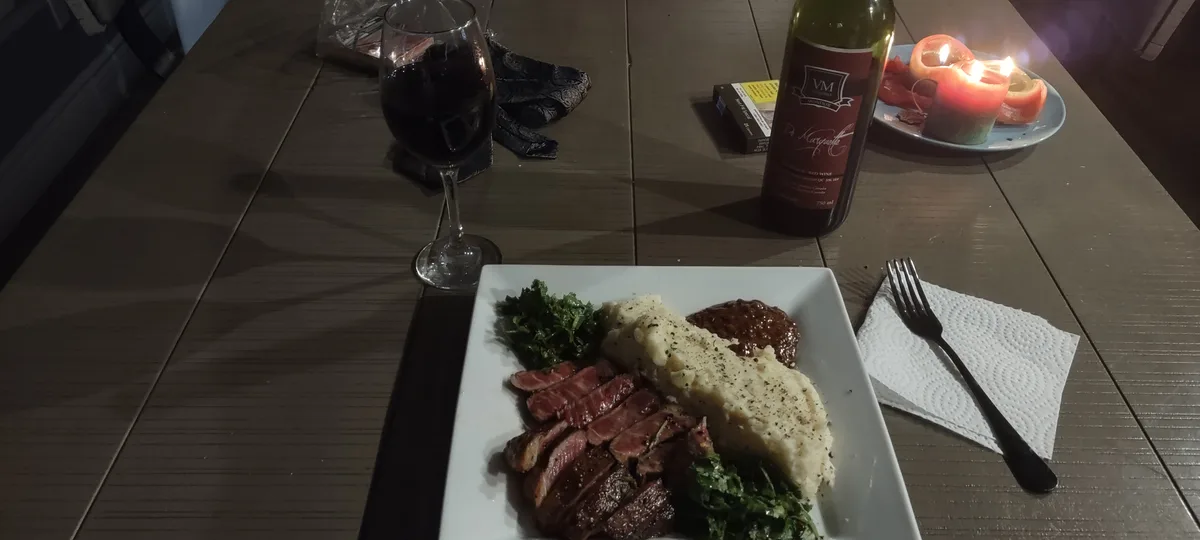 A dimly lit wooden table features a plated meal of sliced steak, mashed potatoes, and greens, alongside a glass and bottle…