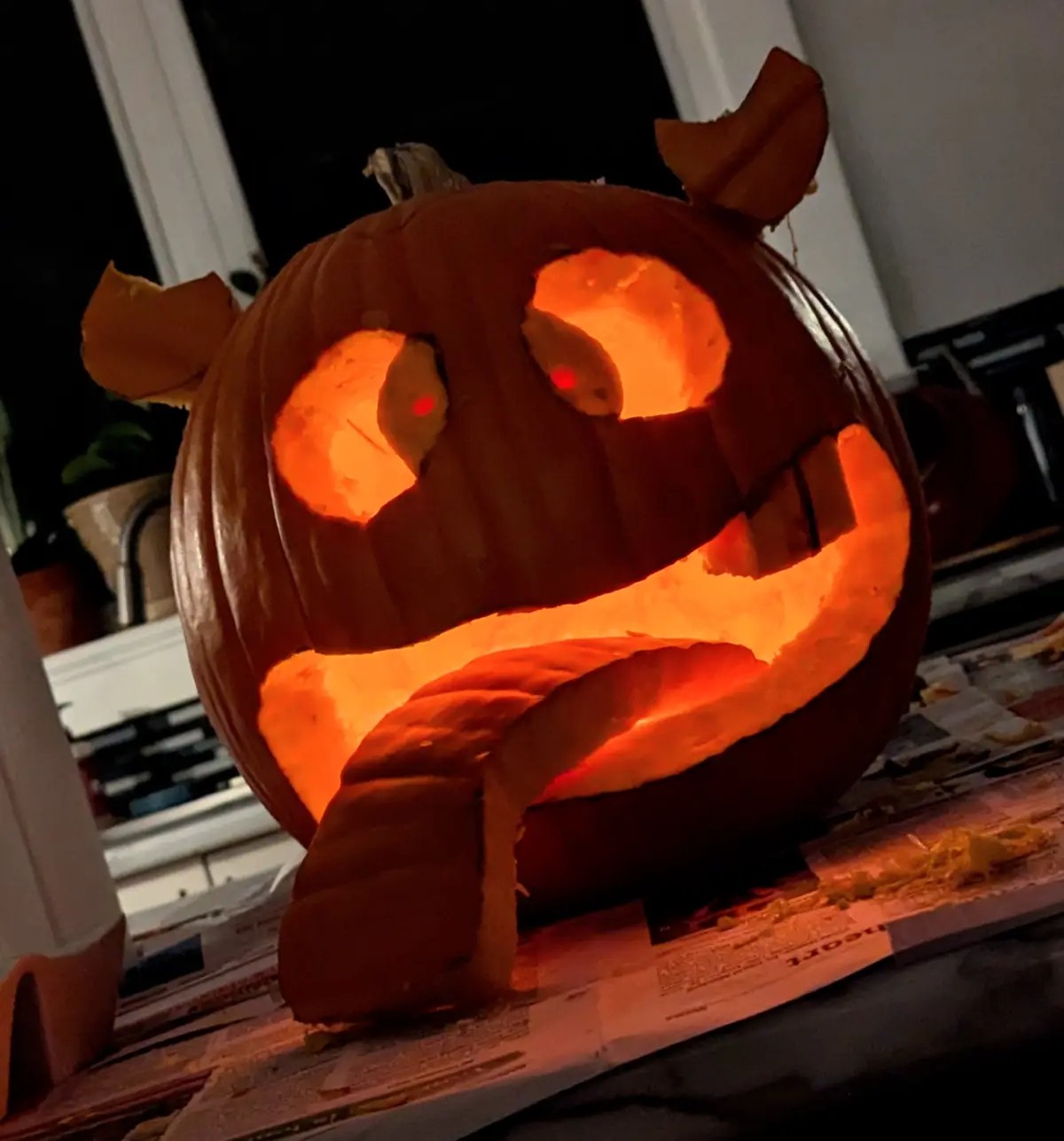 A carved Halloween pumpkin with glowing orange eyes and a wide, toothy grin sits on a newspaper-covered surface, illuminated…