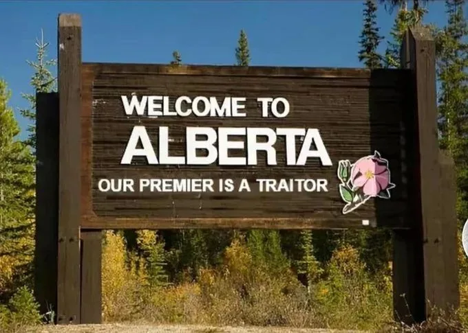 A wooden welcome sign for Alberta, Canada, with white text reading 'WELCOME TO ALBERTA' and below it 'OUR PREMIER IS A…