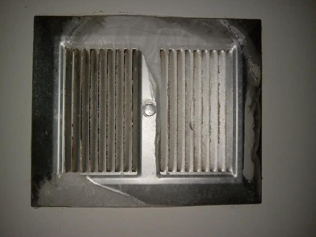 A close-up photo shows a rectangular, metallic vent with vertical slats, mounted on a wall. The vent is visibly dirty, with…