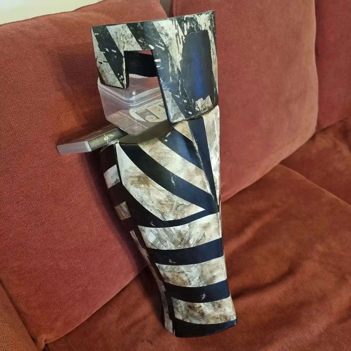 A weathered, black-and-white Union Jack-patterned leg brace or splint rests upright on a rust-colored fabric couch. On top…