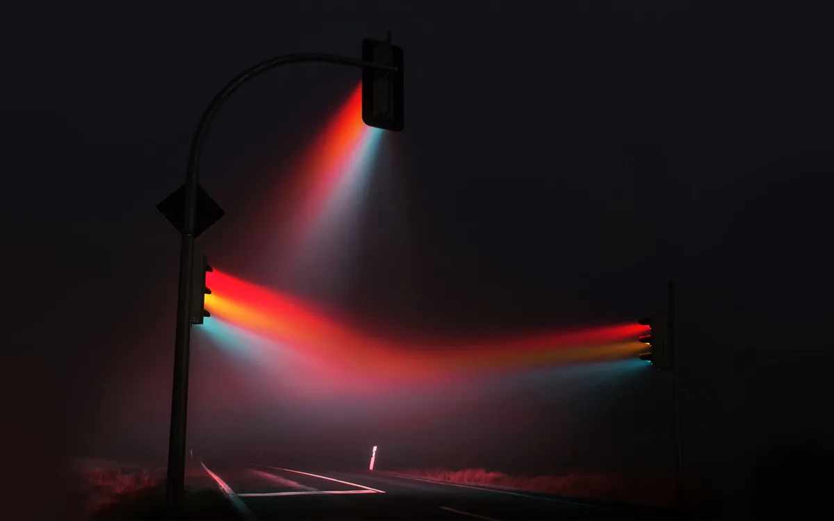 A nighttime photo shows two traffic lights on poles casting vivid beams of red, yellow, and green light across a foggy road…