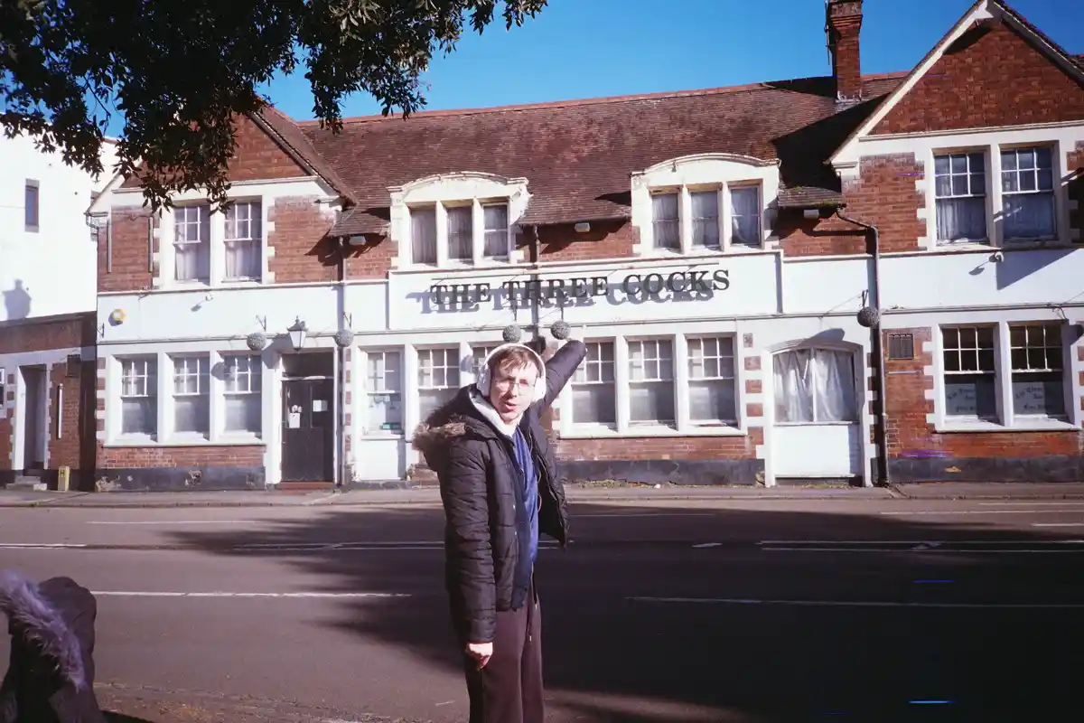 A man wearing headphones and a black puffer jacket stands in front of a brick pub named 'THE THREE COCKS', pointing upward…